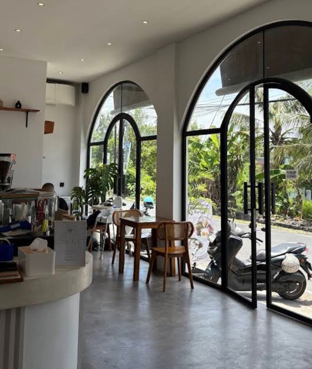 Inside Kopiten: tall arched black-framed windows over a concrete floor, rattan chairs and a teak table, the white fluted counter on the left